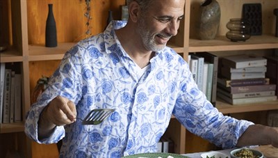 Yotam Ottolenghi in a blue patterned shirt holding a spatula, standing in front of a brown bookcase. Photo