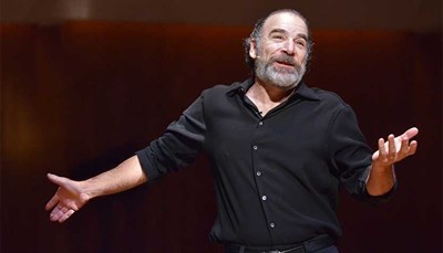 Mandy Patinkin in a black shirt, standing in front of a black background. Photo