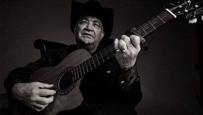 Black and white photo of Eliades Ochoa sitting in a chair with a guitar. Photo