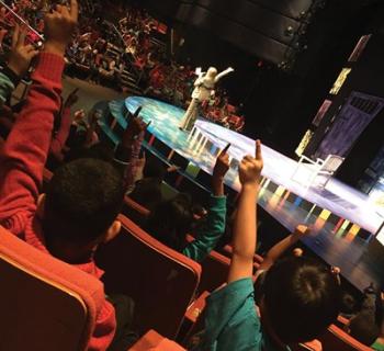 Children raising hands in a theatre. There is an adult on stage.  Photo